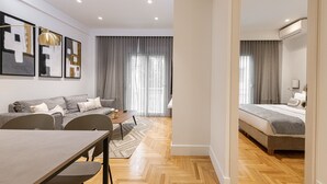 Ett modernt vardagsrum med en soffa, ett matbord och stolar. Det finns ett stort fönster med gardiner, en luftkonditioneringsanläggning och en säng i det intilliggande rummet.