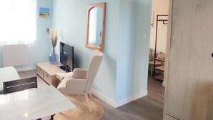Una sala de estar moderna con un sofá blanco, un sillón color beige, una mesita con un jarrón, un televisor y un espejo en la pared.