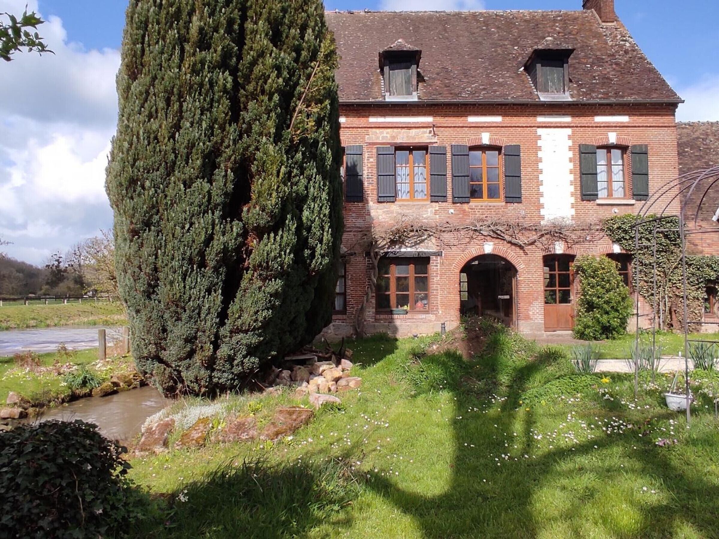 A traditional brick house with a large coniferous tree, a stone pathway, and a small stream.
