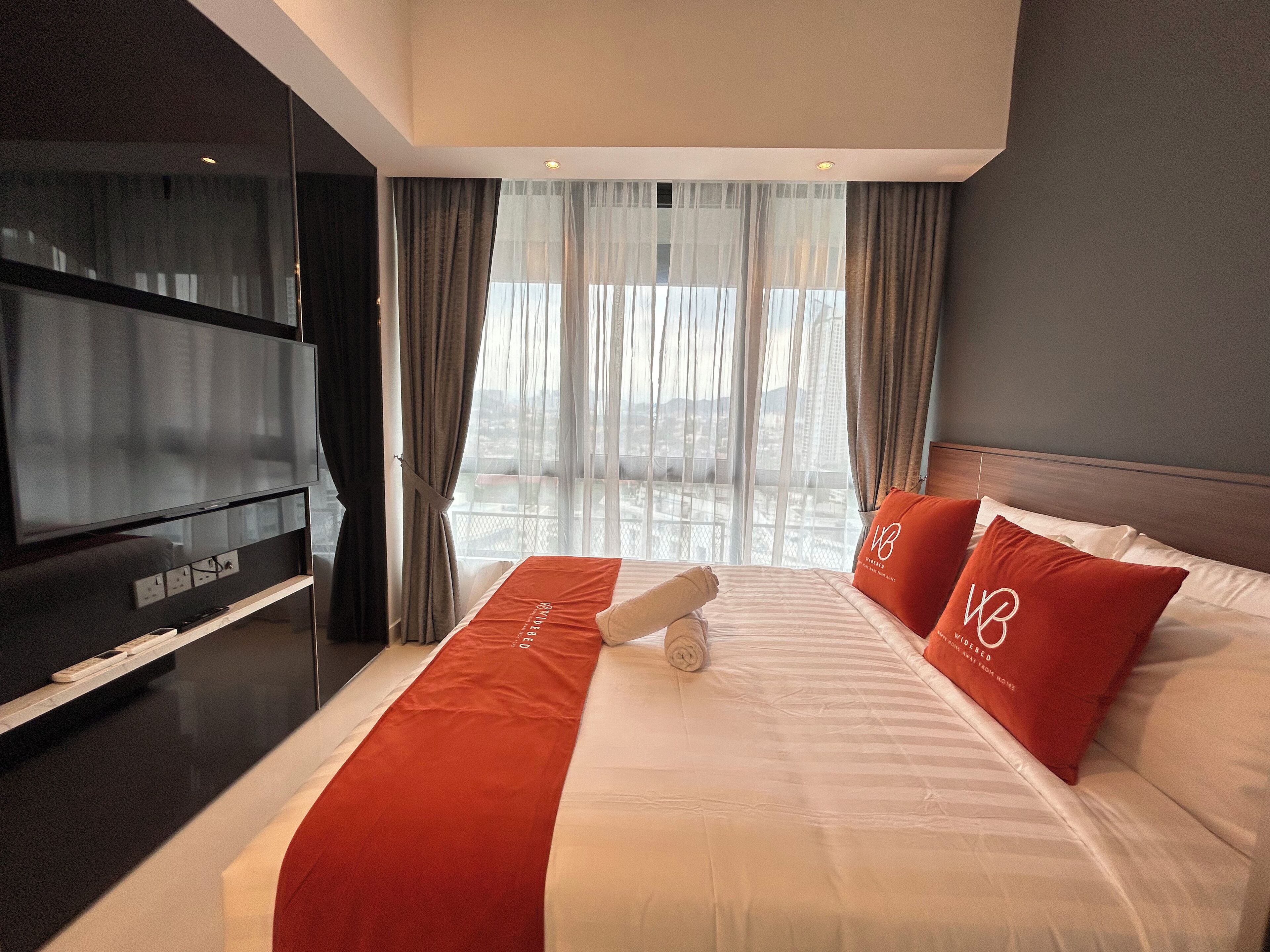 A hotel room with a large bed, a flat-screen TV, and a view of the city through sheer curtains.