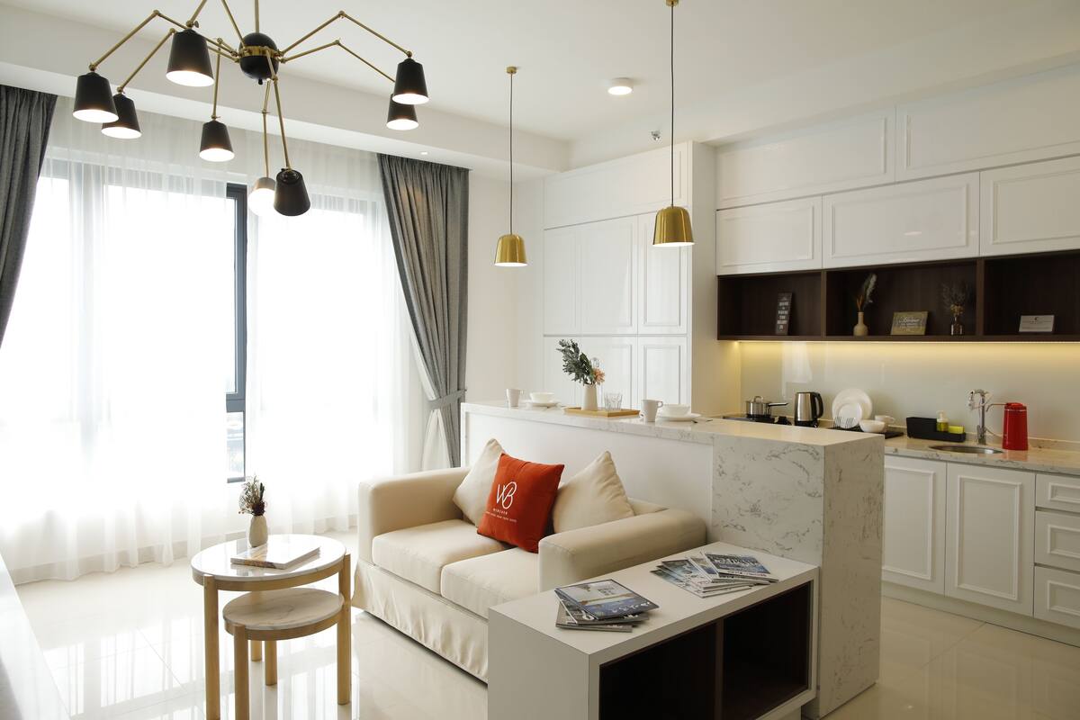 A modern living room with a sofa, a coffee table, and a dining area. There is a kitchen with white cabinets and a marble countertop.