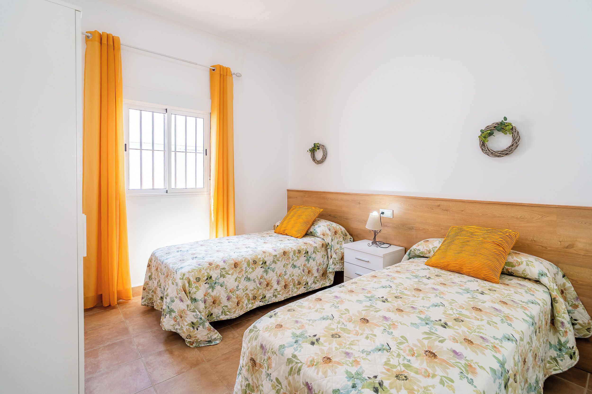 A bedroom with two beds, a nightstand, a window with curtains, and a wall-mounted light.