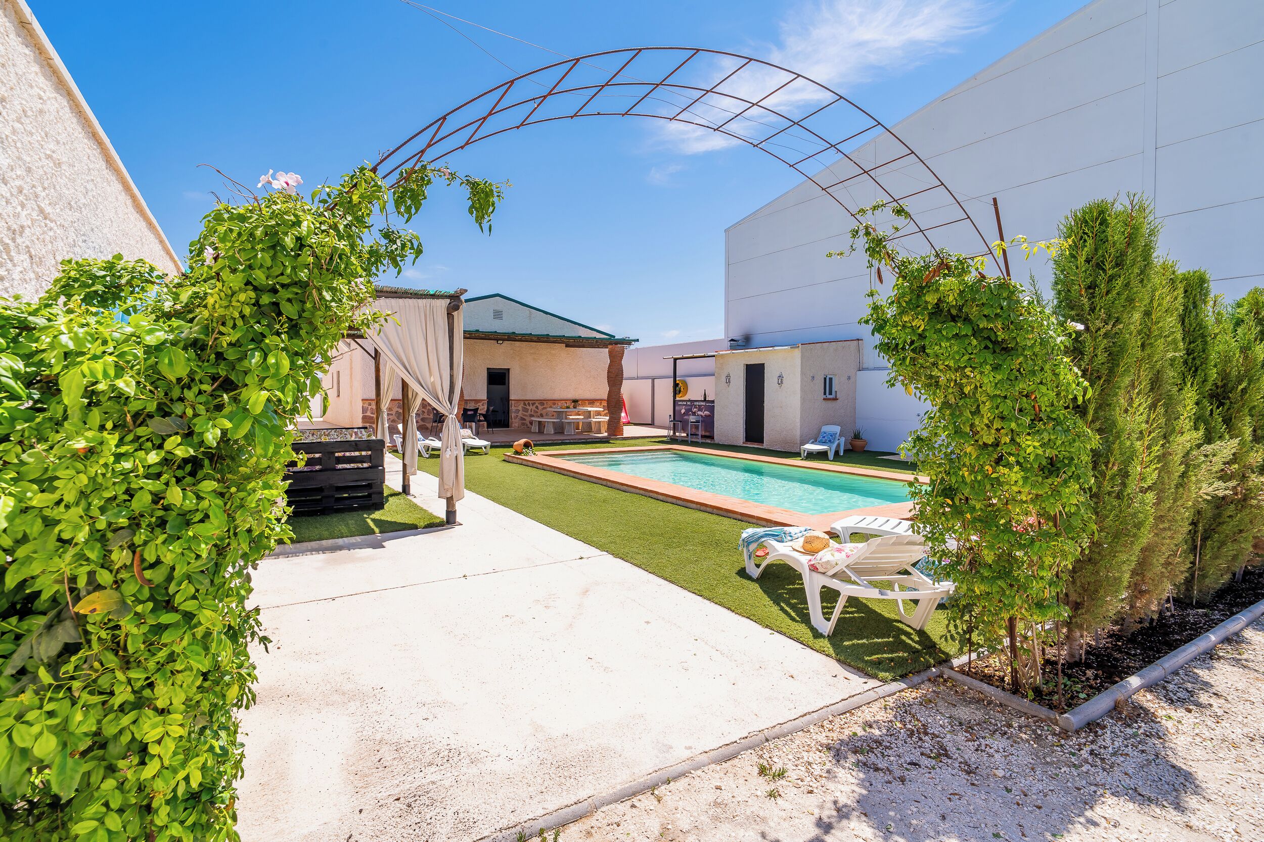 A pool area with a clear sky, a pergola, and a building with a green roof.