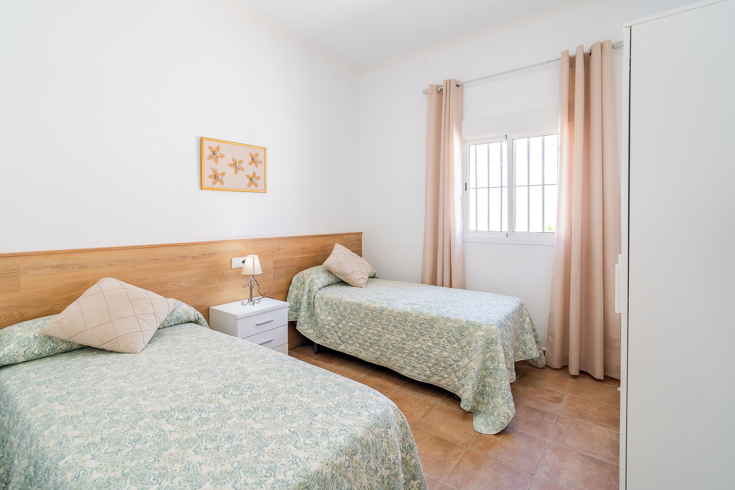 A bedroom with two beds, a nightstand, a lamp, a window with curtains, and a wall-mounted picture.