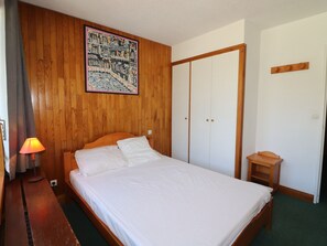 A bedroom with a wooden headboard, a bed with white bedding, a nightstand with a lamp, a wooden wardrobe, and a small wooden table. - Functional apartment with balcony, near slopes, pets allowed (Tignes)