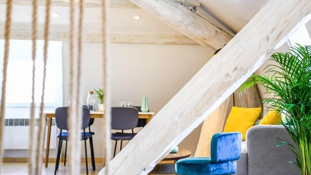 Ein modernes Interieur mit freiliegenden Holzbalken, einem Essbereich mit Stühlen und Tisch sowie einer Sitzecke mit Pflanze.