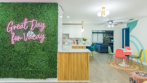 Una sala moderna con una pared verde, un letrero de neón, una isla de madera y una zona de comedor con sillas coloridas.