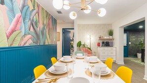 Un comedor con una mesa larga dispuesta para seis personas, rodeada de sillas amarillas. La habitación presenta un mural llamativo en la pared, una lámpara de techo moderna y un área de cocina visible a través de un umbral abierto.