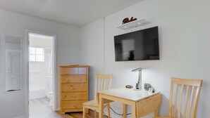 Une petite pièce bien éclairée, avec une commode en bois, un bureau avec une chaise, un téléviseur fixé au mur et une porte donnant sur une autre pièce.