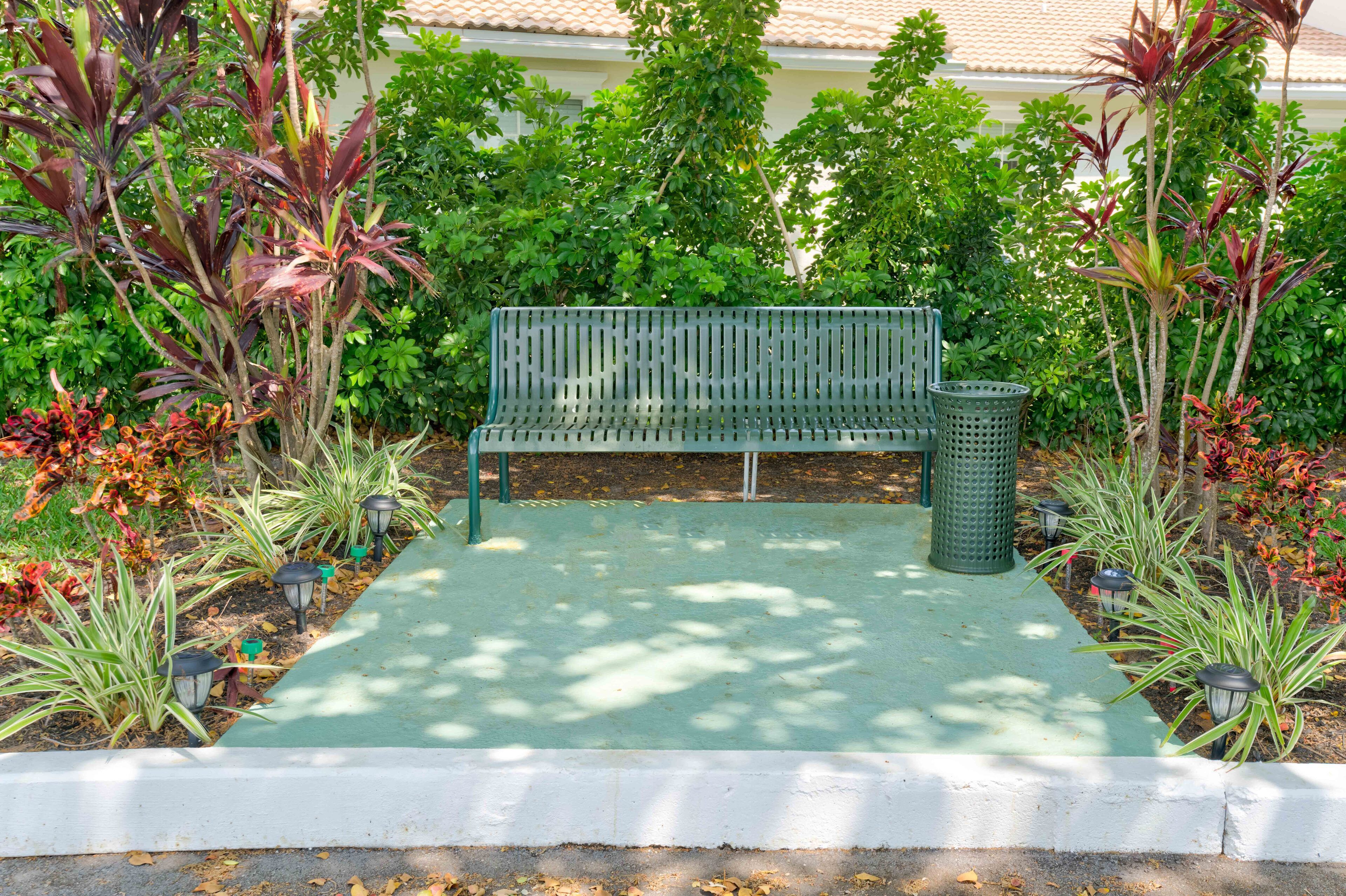 Un banco verde en un jardín con diversas plantas y un cubo de basura.