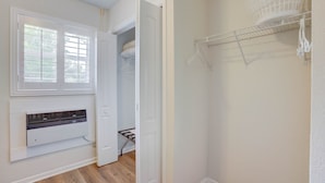 Une pièce avec un radiateur blanc, une fenêtre avec des stores, une porte donnant sur un placard et une étagère blanche fixée au mur, sur laquelle se trouve un panier.