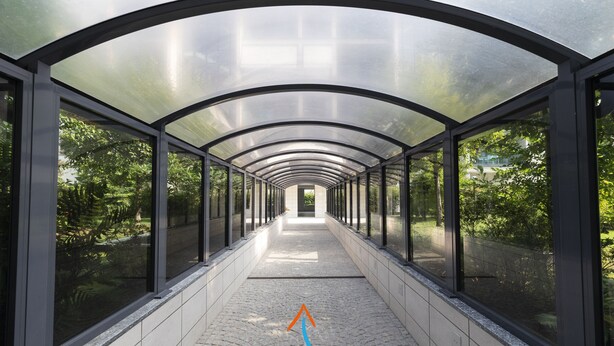 A covered walkway with glass walls and a tiled floor, surrounded by greenery.