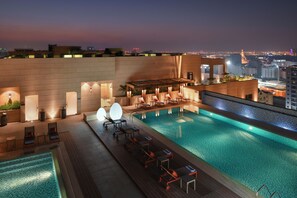 Un espace piscine aménagé sur le toit, avec des chaises longues et un bar, offrant une vue sur le paysage urbain au crépuscule.