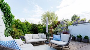 Een terras buiten met rieten meubels, een witte muur en potplanten.