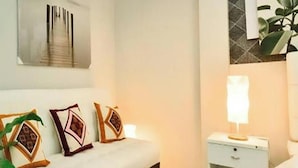 Een moderne slaapkamer met een wit bed, decoratieve kussens, een nachtkastje met een lamp en een aan de muur hangend kunstwerk.