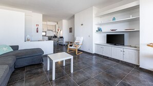 Ein modernes Wohnzimmer mit grauem Sofa, einem weißen Couchtisch und einem Holzstuhl mit farbenfroher Sitzkissen. Ein Flachbildfernseher ist an einem weißen Schrank befestigt, und im Hintergrund ist eine Küchenzeile zu sehen.