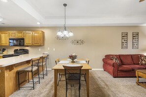 La cuisine est équipée de meubles en bois, d’un réfrigérateur en acier inoxydable et d’un four encastré. On y trouve également un coin repas avec une table en bois et quatre tabourets de bar. Le salon est agrémenté d’un canapé rouge et d’une table basse en bois.