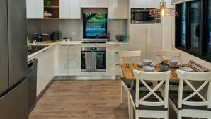 A modern kitchen with white cabinets, a built-in oven, and a dining area with white chairs and a wooden table. - ZEN AT ROSEWOOD - 2BR Guesthouse & Pool (Leanyer)