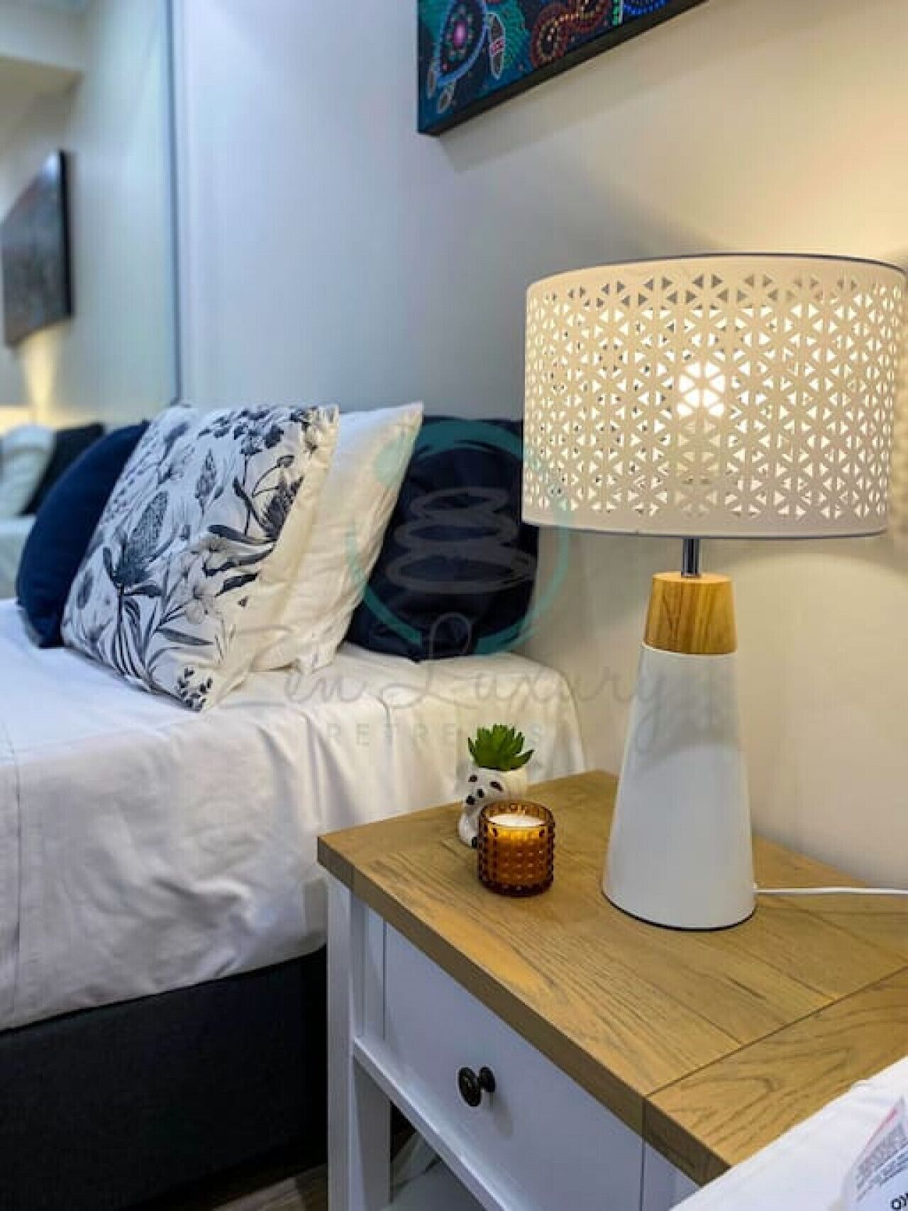 Cama bem arrumada com almofadas estampadas, mesa de cabeceira com abajur decorativo, uma pequena planta e uma gaveta.