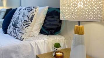 Cama bem arrumada com almofadas estampadas, mesa de cabeceira com abajur decorativo, uma pequena planta e uma gaveta.