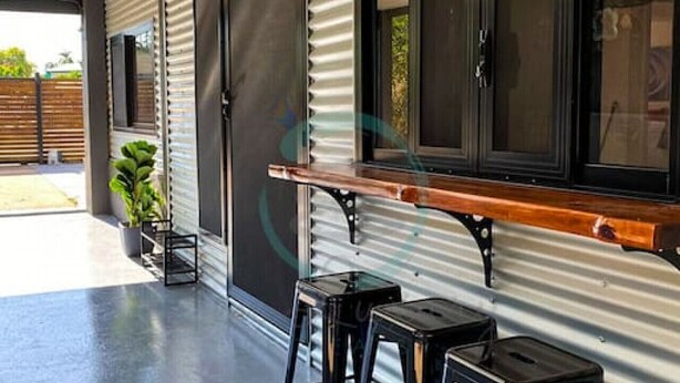 A modern outdoor seating area with metal-framed tables and black stools, a corrugated metal wall, and a wooden bench. - ZEN AT ROSEWOOD - 2BR Guesthouse & Pool (Leanyer)
