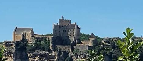 A historic stone building with a prominent tower, situated on a rocky hill with lush greenery in the foreground.