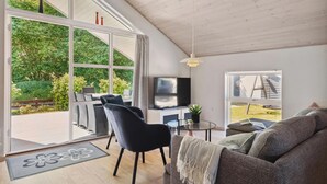 Un salon moderne doté d’une grande porte-fenêtre coulissante donnant sur un jardin, d’un téléviseur à écran plat, d’un canapé et d’un coin repas avec des chaises.
