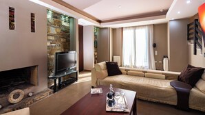 A modern living room with a stone fireplace, a flat-screen TV, a sofa, and a coffee table with a tray. - Villa Astravi with Private Pool (Ιαλυσός)