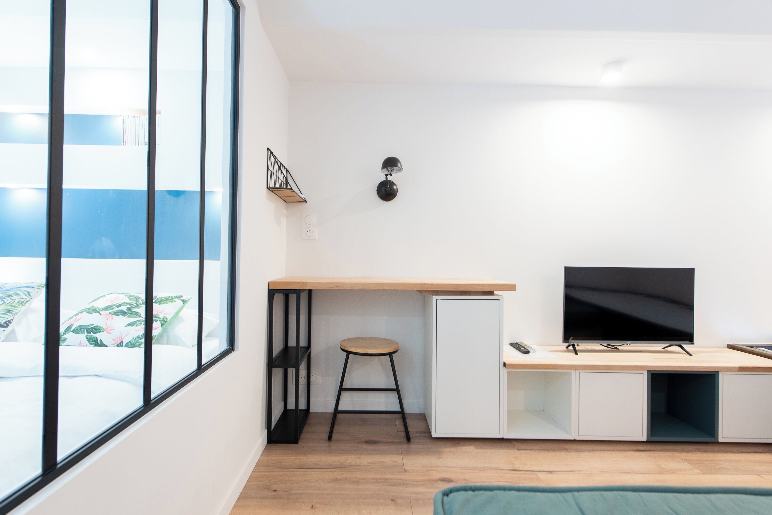 Een moderne kamer met een groot raam, een bureau met een televisie, een kruk en een boekenplank.