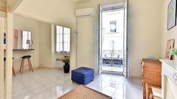 A bright living room with a large window, a wicker ottoman, a blue stool, and a wicker chair.