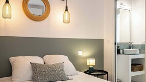 Una cama bien hecha con dos almohadas, una lámpara de noche y un espejo en la pared.