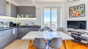 A modern kitchen with grey cabinets, a white dining table, and a large window. - Le Sinatra splendide vue mer panoramique et balcon (Cannes)