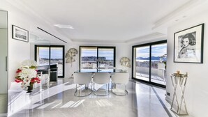 A modern, well-lit room with a dining area featuring a glass table and white chairs, large windows offering a city view, and a vase of flowers on the floor. - L'Horizon avec somptueuse terrasse vue mer (Cannes)