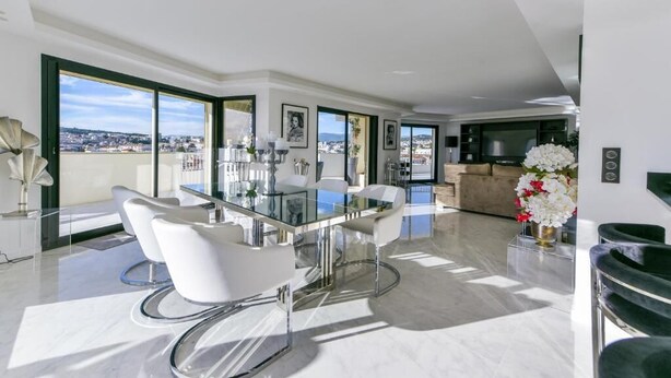A modern living room with a glass dining table, white chairs, and a sofa. There are large windows with a city view, a flat-screen TV, and a decorative vase with flowers. - L'Horizon avec somptueuse terrasse vue mer (Cannes)