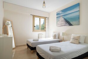 A hotel room with two beds, a window, a painting of a pier, a desk with a lamp, and a radiator.