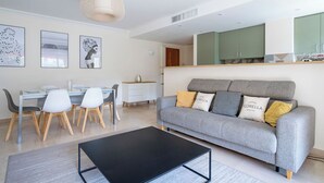 Een moderne woonkamer met een grijze bank, een zwarte salontafel en een eethoek met een glazen tafel en stoelen.