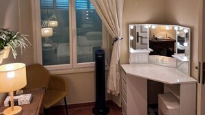 A hotel room with a large window, a vanity with a mirror and lights, a chair, a lamp, and a potted plant.
