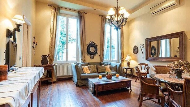 A cozy living room with a sofa, coffee table, and dining table. There are two windows with curtains, a chandelier, and a mirror on the wall.
