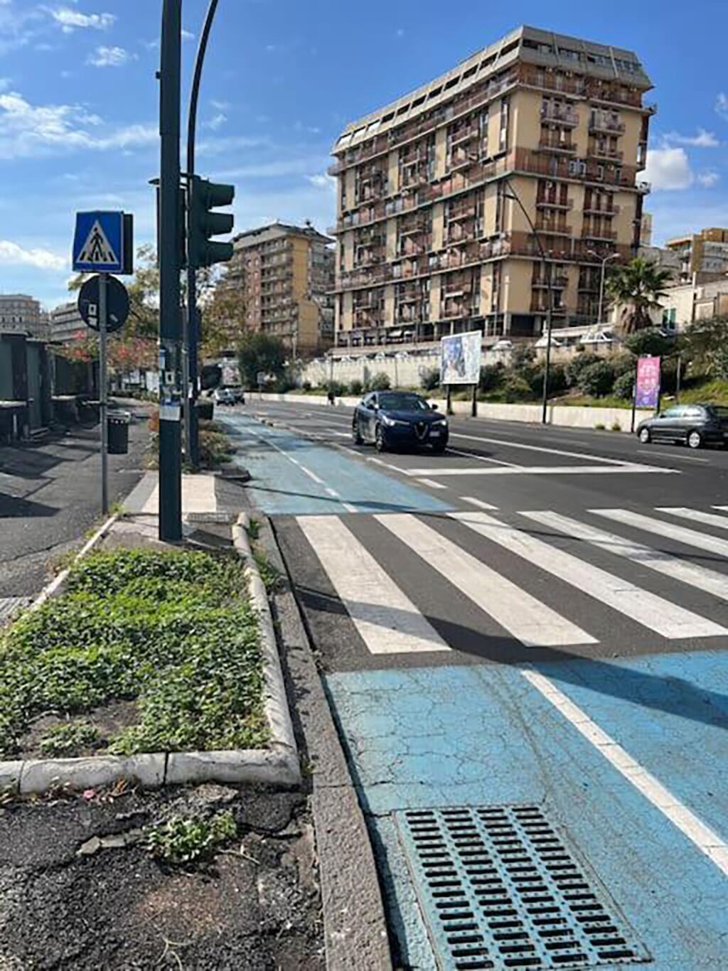 Un passage piéton avec une bande bleue peinte au sol, un feu de signalisation et un immeuble à plusieurs étages en arrière-plan.
