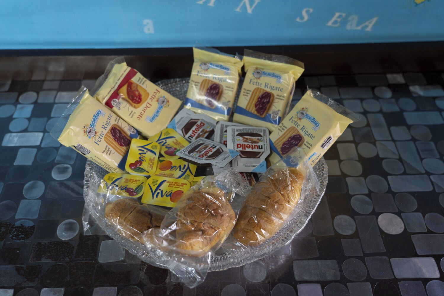 Un cuenco de vidrio con diversos productos alimenticios envasados y dos barras de pan sobre una superficie de baldosas.