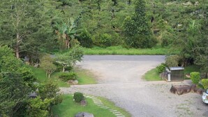 Uma entrada com portão no final de um caminho de cascalho, cercada por vegetação exuberante e diversas árvores.