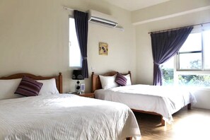 A hotel room with two beds, a nightstand, an air conditioning unit, and a window with curtains.