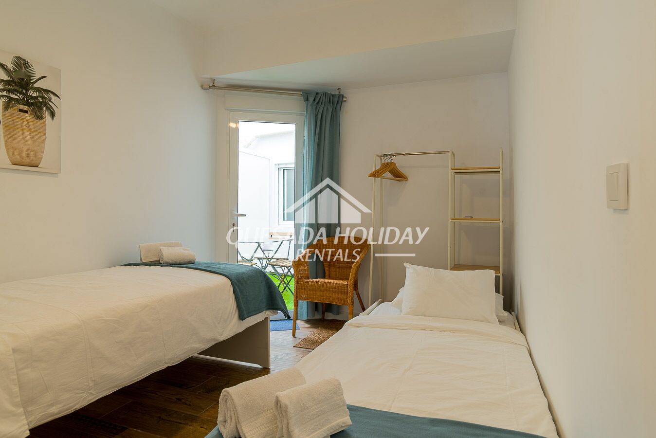Une chambre d’hôtel avec deux lits simples, une petite table, une chaise, un porte-manteau et une grande fenêtre avec des rideaux.