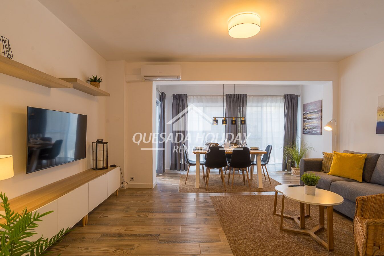 A modern living room with a flat-screen TV, a dining area with a table and chairs, and a sofa with a yellow cushion.