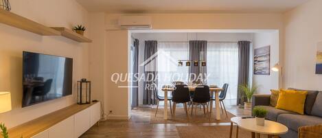 A modern living room with a flat-screen TV, a dining area with a table and chairs, and a sofa with a yellow cushion.