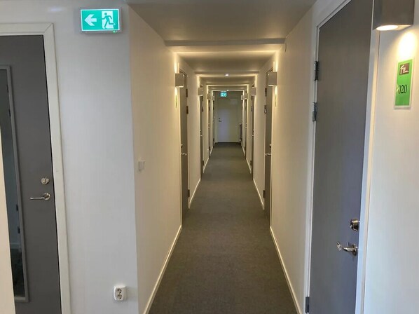 A hotel corridor with multiple doors, emergency exit signs, and a carpeted floor. - Micro Hotel Apartments (Upplands Vasby)