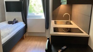 Habitación pequeña con cama, una kitchenette compacta con lavabo y estufa, un televisor montado en la pared y una ventana con cortinas.