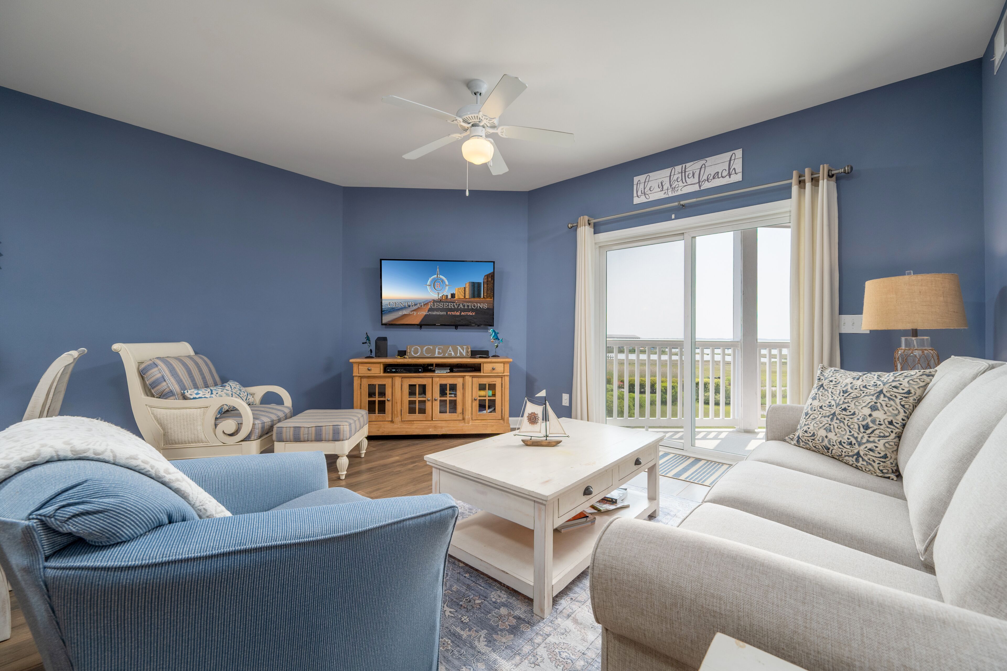 A living room with a blue wall, a white ceiling fan, a television, a wooden cabinet, and a view of the outdoors.
