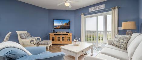 A living room with a blue wall, a white ceiling fan, a television, a wooden cabinet, and a view of the outdoors.