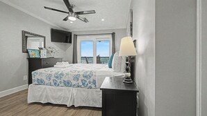 Une chambre à coucher comprenant un lit, un bureau, une chaise, un ventilateur de plafond, une télévision et une fenêtre donnant sur un beau paysage.